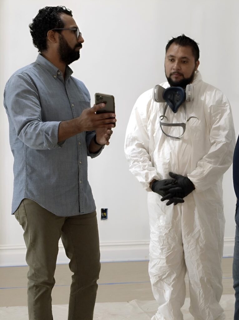Man in a blue shirt holding a phone beside a worker in a white protective suit, respirator and black gloves