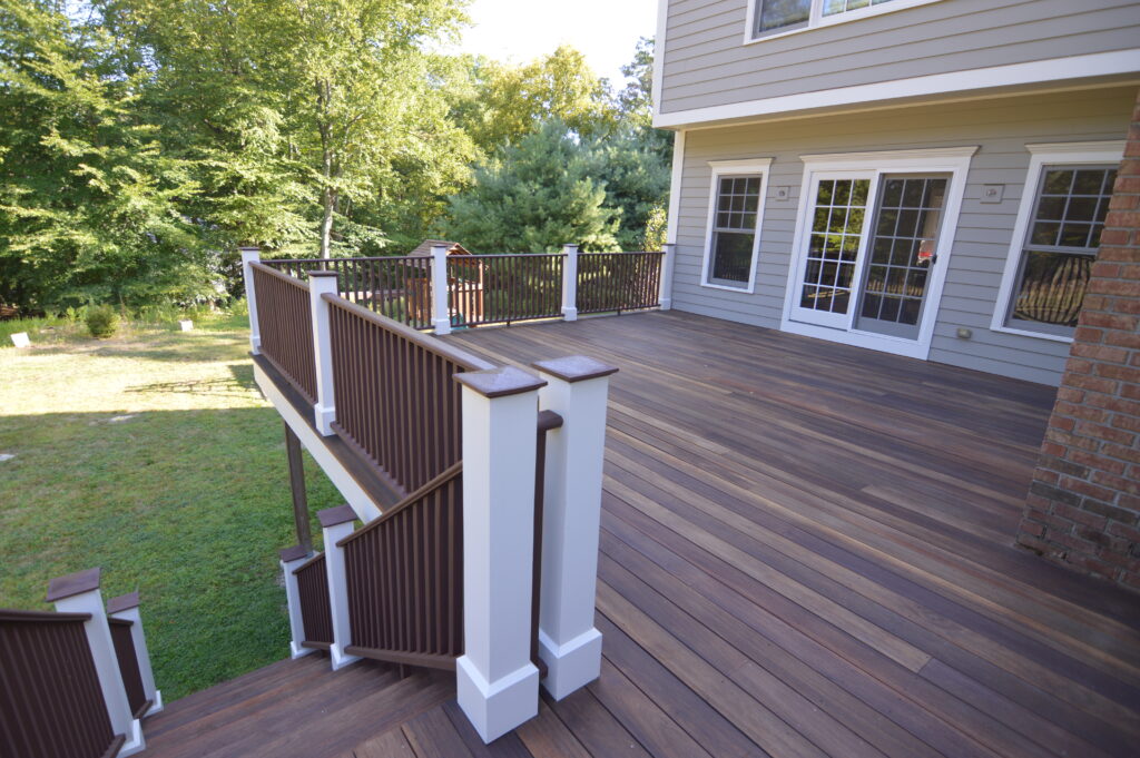 Dark stained wood deck with white post sleeves, brown railings and steps down to the lawn beside a gray sided house