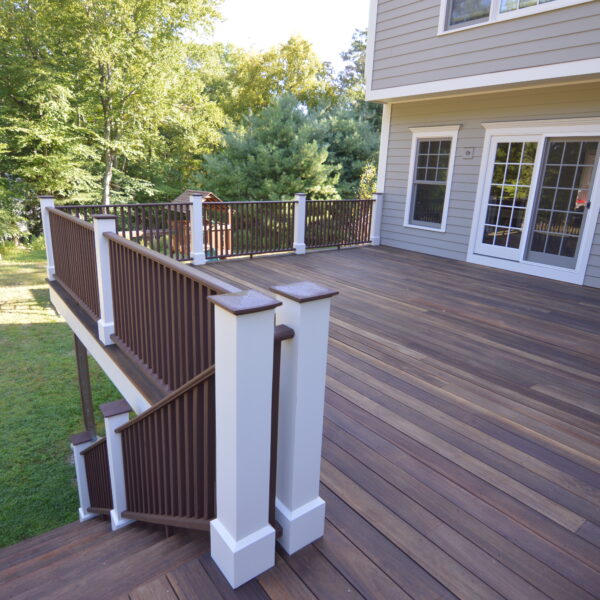 Dark stained wood deck with white post sleeves, brown railings and steps down to the lawn beside a gray sided house