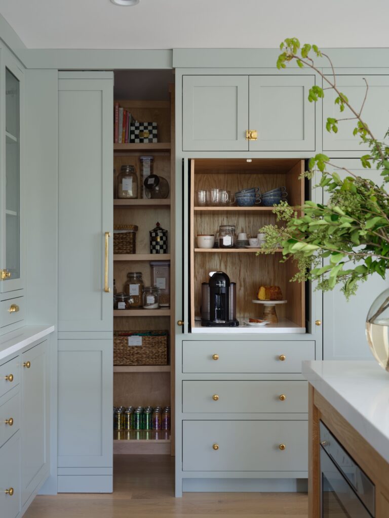 Sage green kitchen cabinets with brass pulls and open pantry doors showing wood shelves and a coffee station