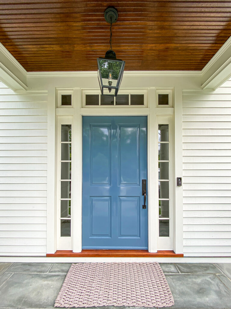 Glossy blue paneled front door with glass sidelights and a transom under a stained wood porch ceiling