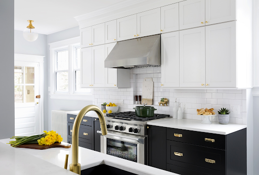 Kitchen with white upper cabinets, black lower cabinets with brass pulls, a stainless range hood and a brass faucet