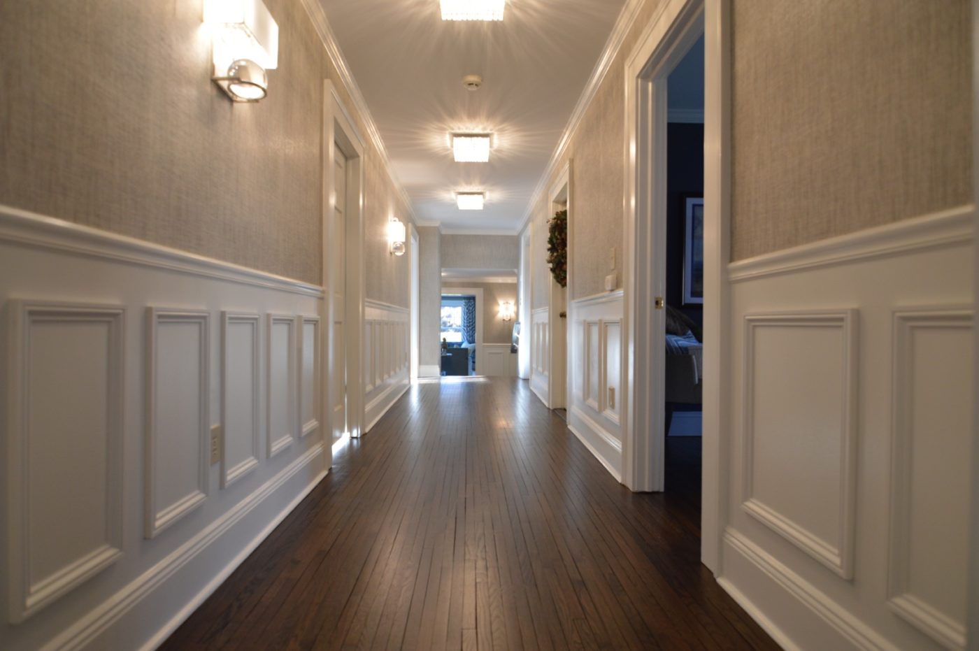 Long hallway with white panel molding wainscoting, textured wallpaper above and dark hardwood floors