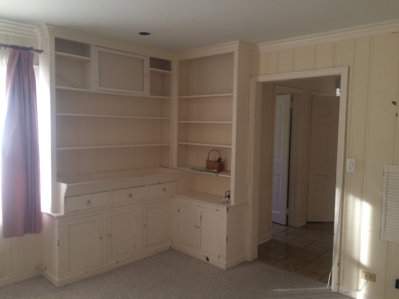 Room with worn cream built-in shelves and cabinets with chipped paint, paneled walls and light carpet