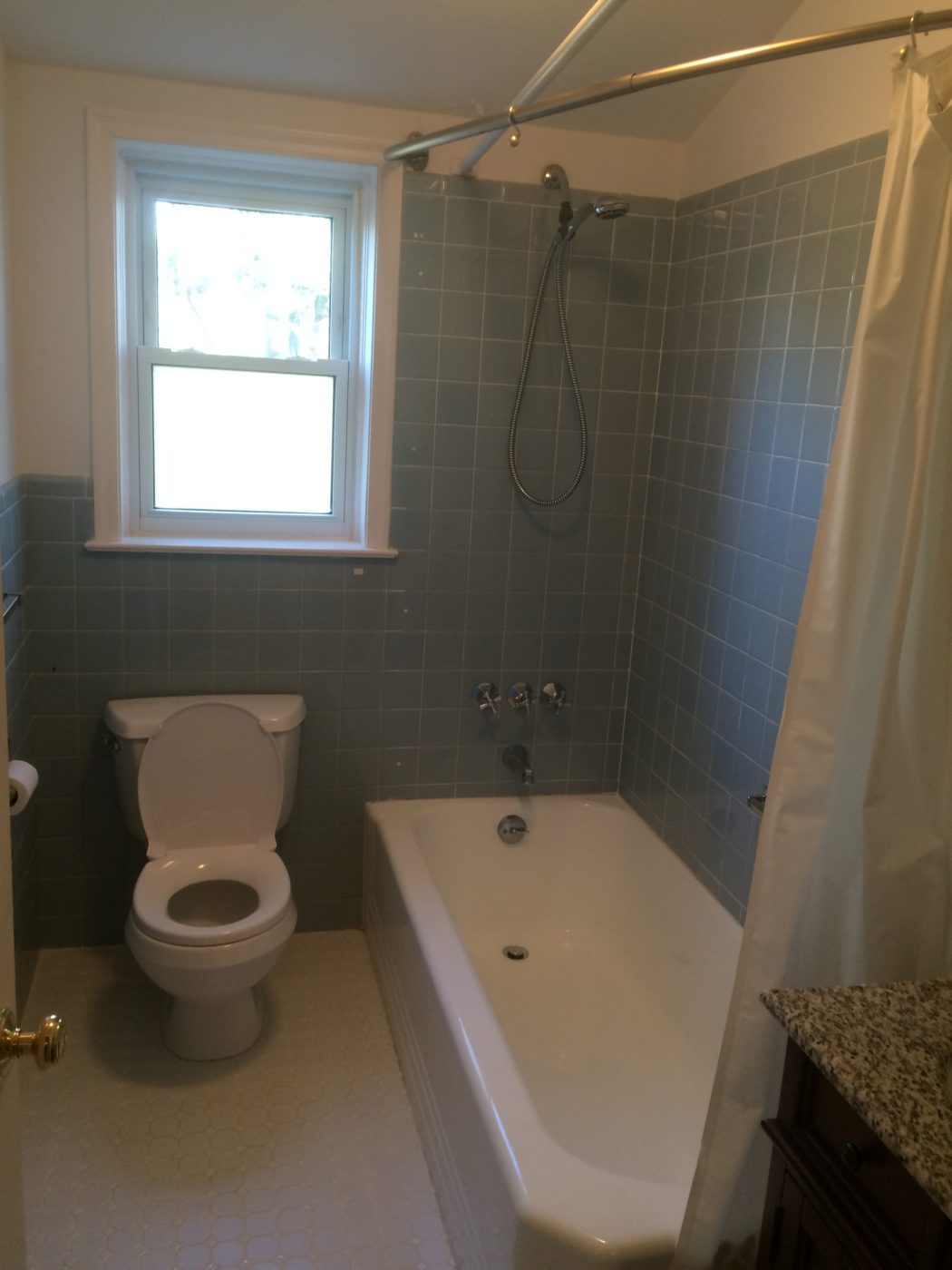 Bathroom with blue-gray wall tile around a white tub, a hand shower, a shower curtain and white hex floor tile