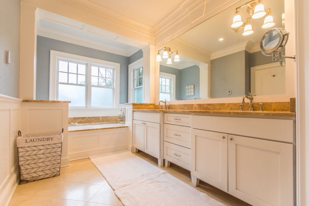 Bathroom with a long white vanity, a gold-toned stone countertop, a soaking tub under the windows and pale blue walls