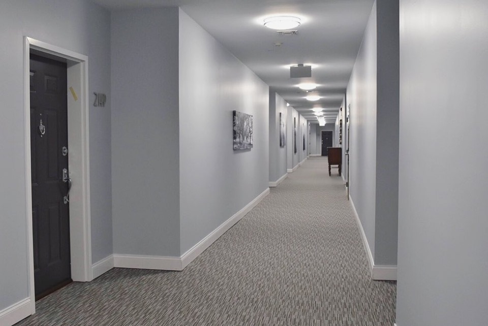 Long apartment building corridor with pale gray walls, white baseboards and a dark unit door