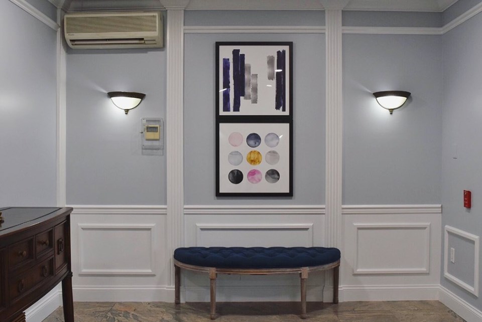 Building lobby with pale blue-gray walls, white wainscoting panels, two wall sconces, framed art and a navy bench