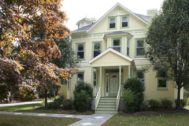 Victorian house painted pale yellow with soft green trim, windows and front door, framed by autumn trees