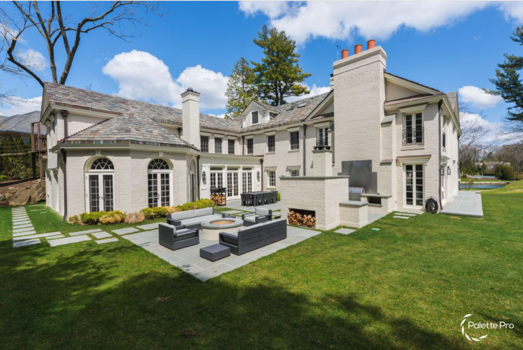 Rear of a large white house with a slate roof, a patio with outdoor sofas, an outdoor fireplace and a green lawn