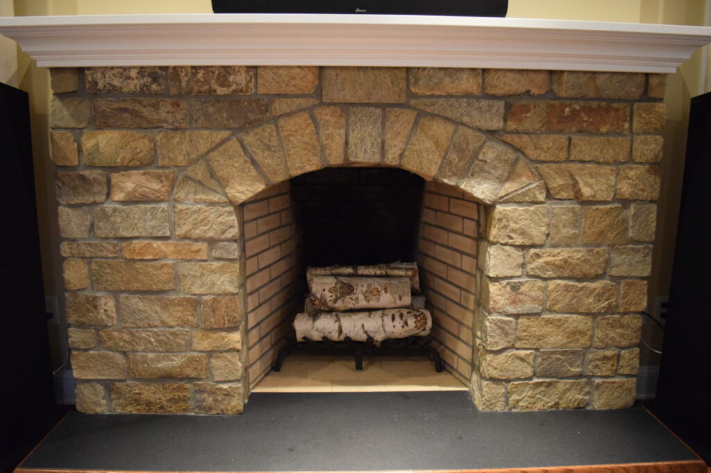 Stone fireplace with an arched opening, a white mantel shelf and birch logs on the grate