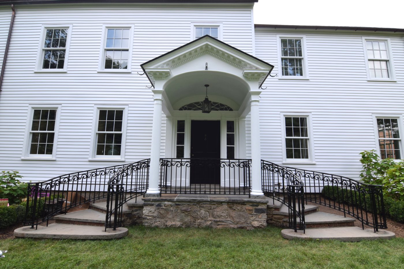 White clapboard farmhouse with a columned front portico, black front door and curved iron stair railings