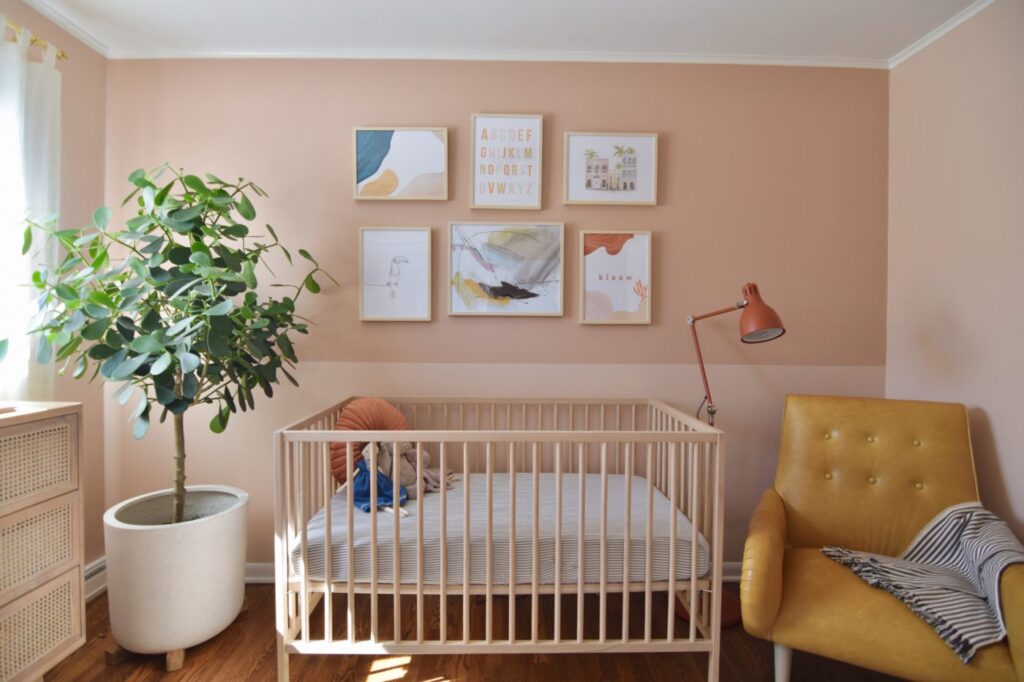 Nursery with blush pink walls, a white crib, framed art prints, a mustard yellow armchair and a potted tree