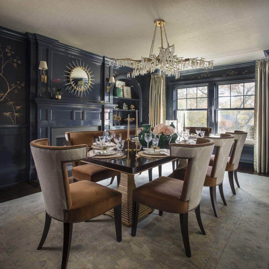 Dining room with glossy navy paneled walls, a gold branch wall covering, a crystal chandelier and cognac velvet chairs
