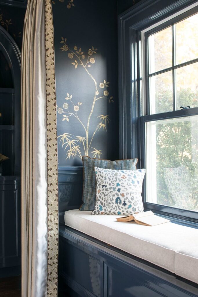 Window seat with a cream cushion and patterned pillows in a glossy navy room with a gold branch wall covering