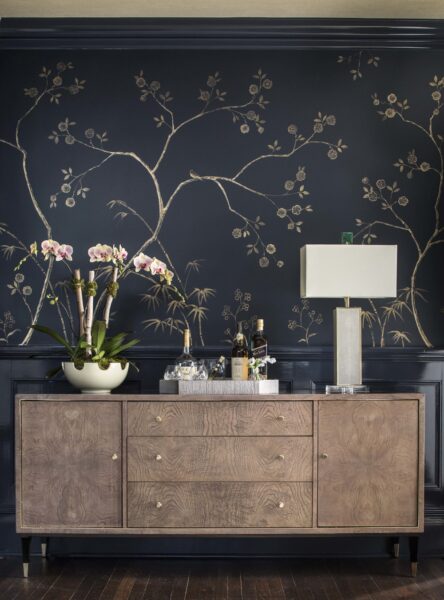 Navy wall covering with a gold branch and blossom pattern above a wood sideboard with orchids, bottles and a lamp