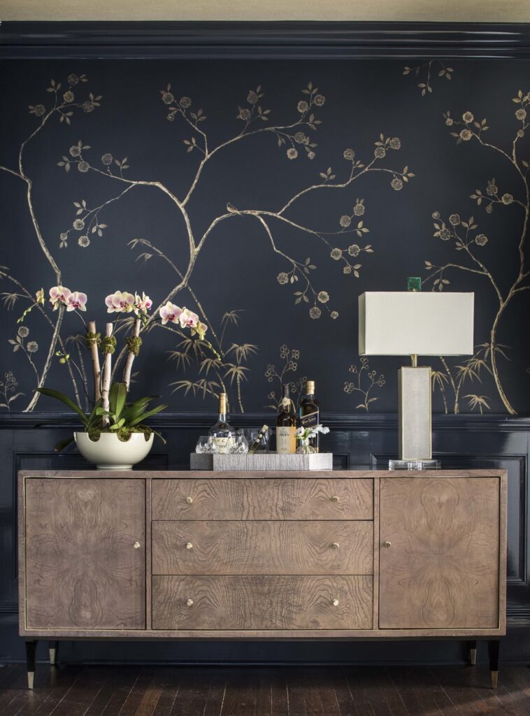 Navy wall covering with a gold branch and blossom pattern above a wood sideboard with orchids, bottles and a lamp