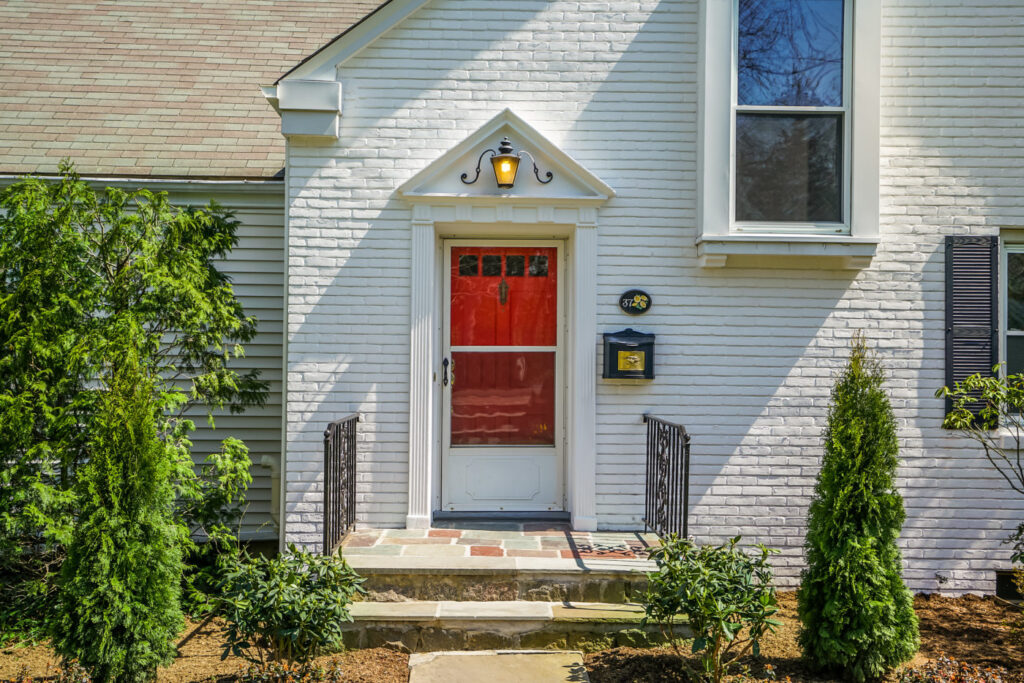 White painted brick house with a red front door, a lantern under a small pediment, dark shutters and evergreens