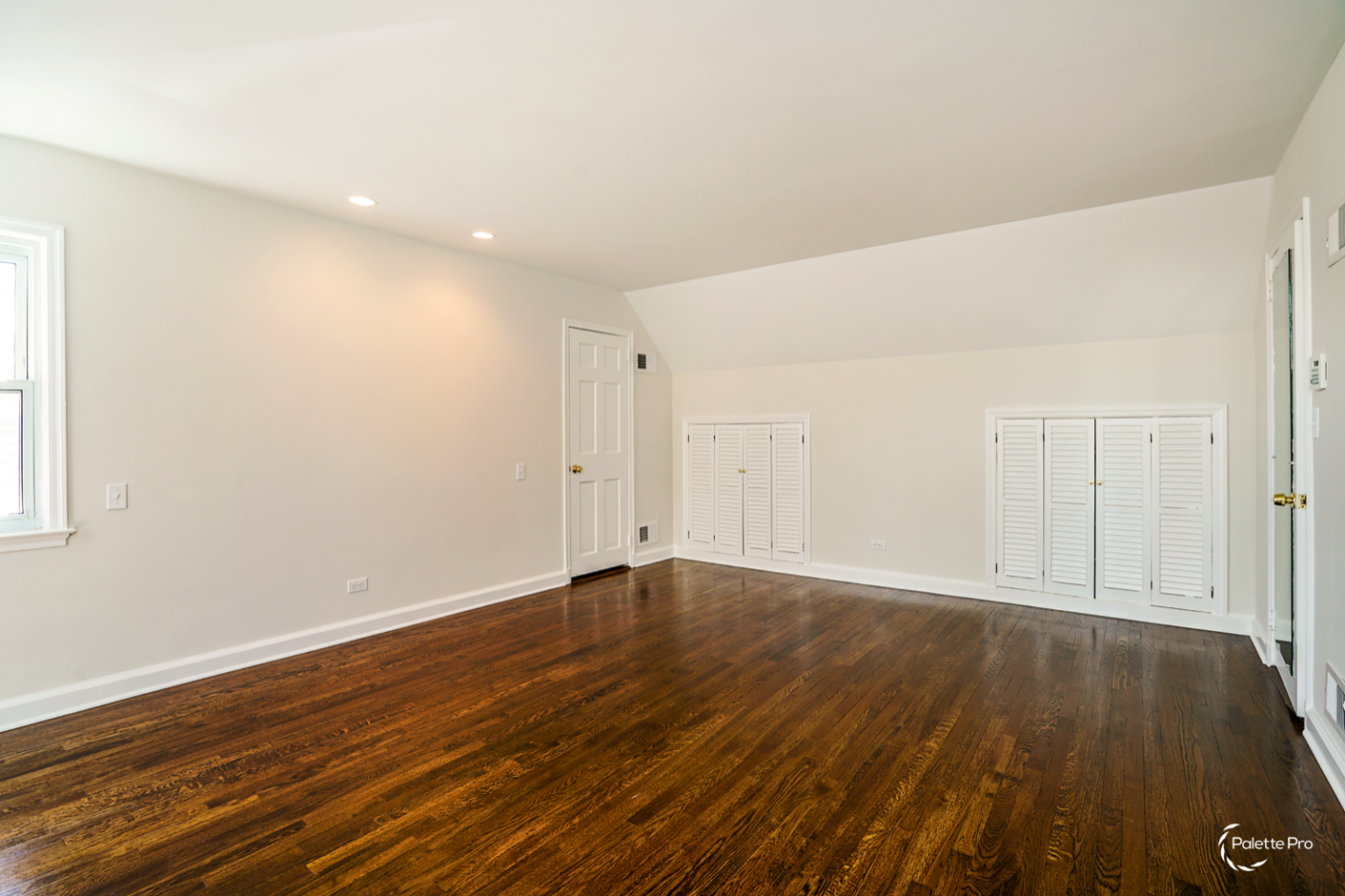 Empty bedroom with refinished dark hardwood floors, off-white walls and white louvered closet doors