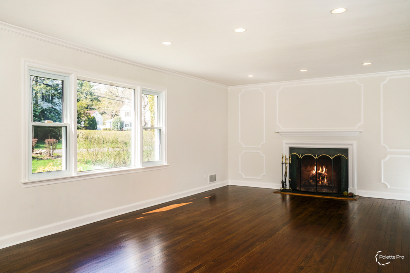 Empty living room with refinished dark hardwood floors, white walls with panel molding and a lit fireplace