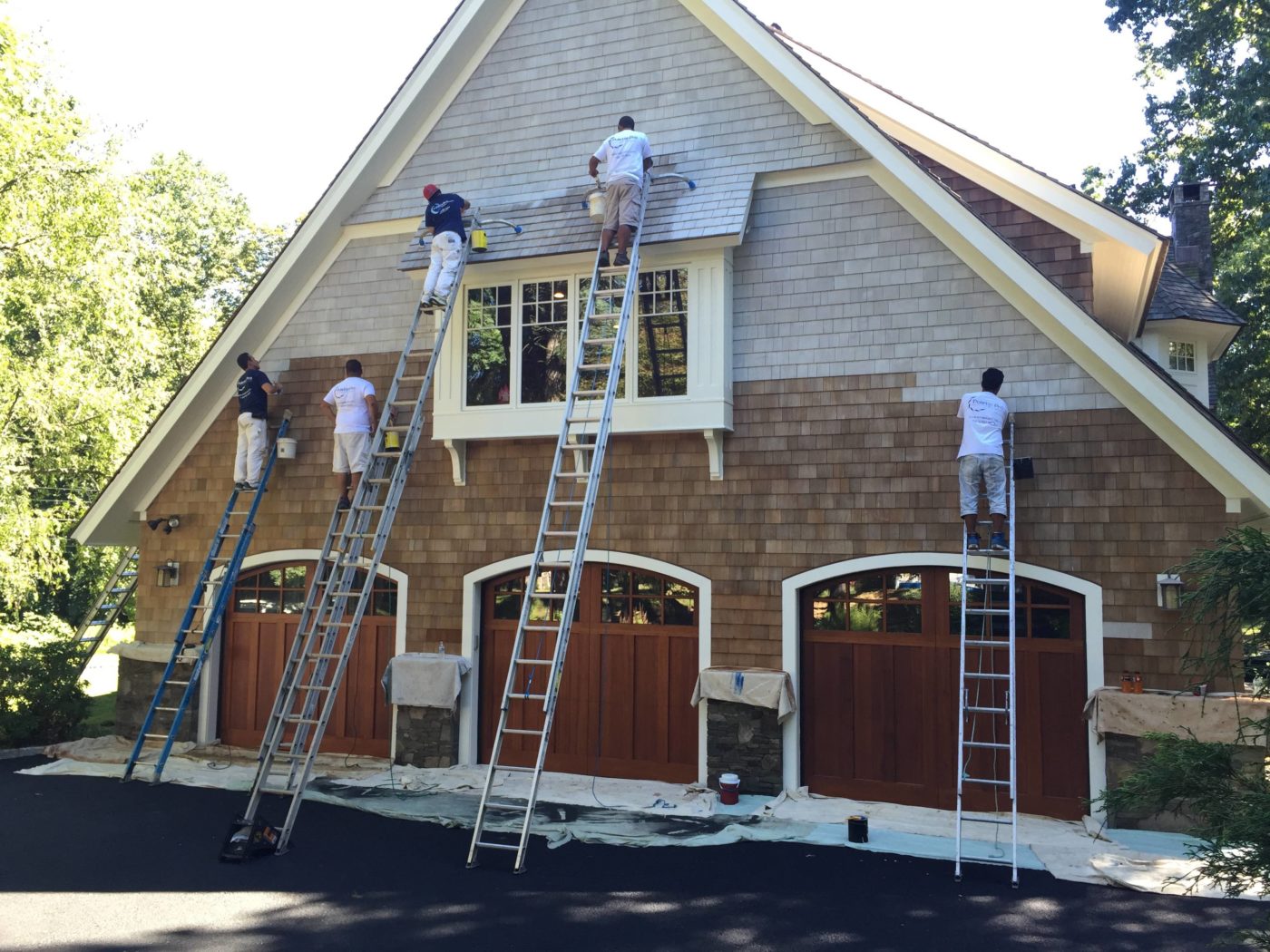 Painters on tall ladders working on the cedar shingle siding of a house above three arched wood garage doors