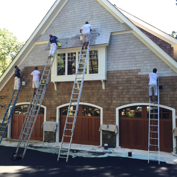 Painters on ladders refinishing cedar shingles in gray around cream trim above three wood carriage garage doors