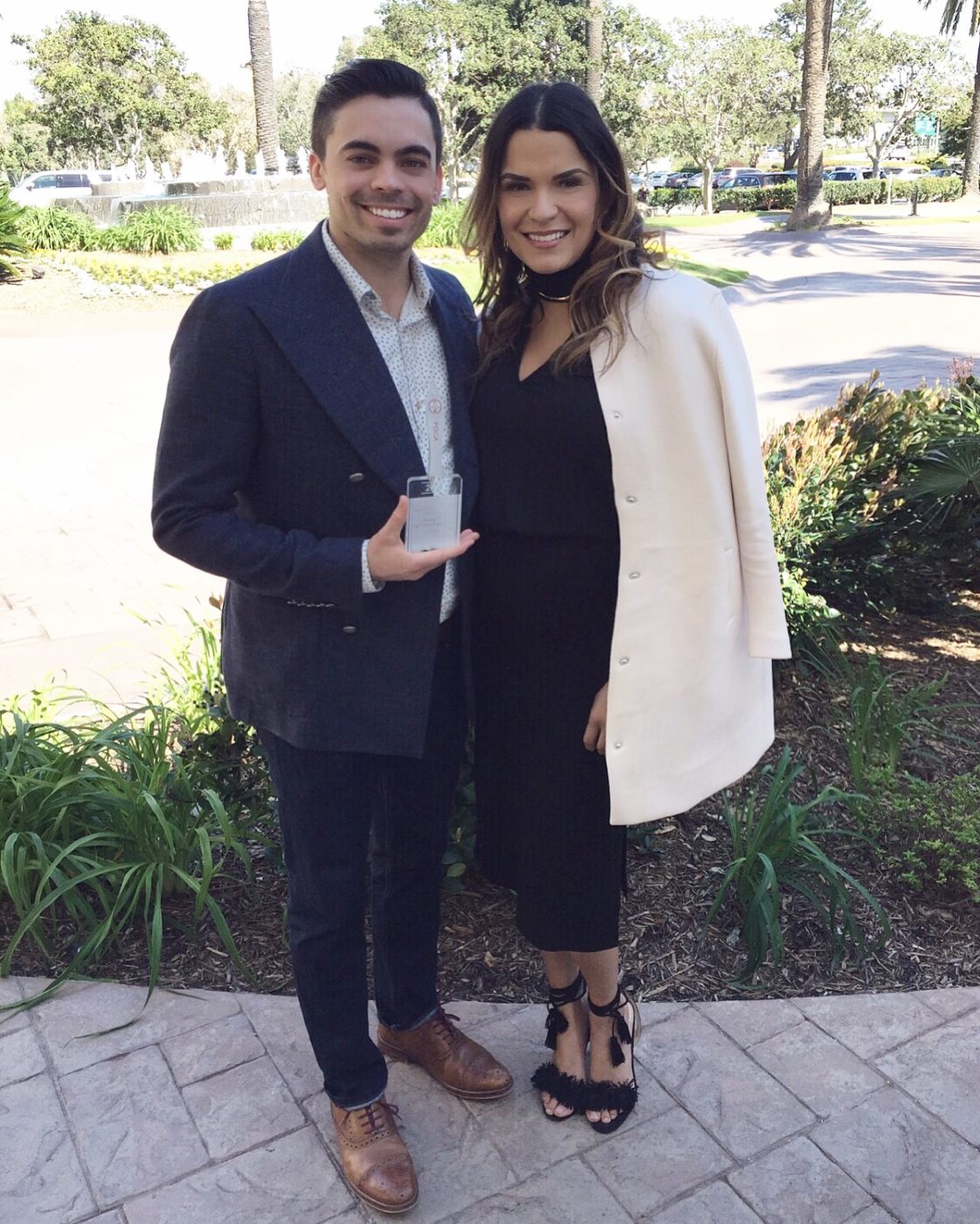 Man and woman posing outdoors on a sunny day, the man holding a small glass award