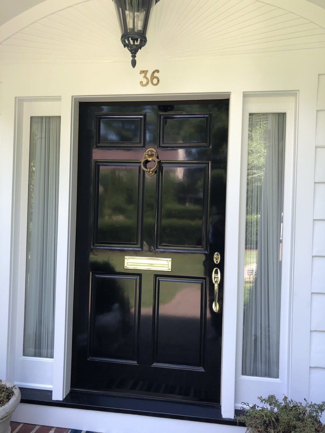 Glossy black paneled front door with a brass knocker and mail slot between white sidelights, under a black lantern
