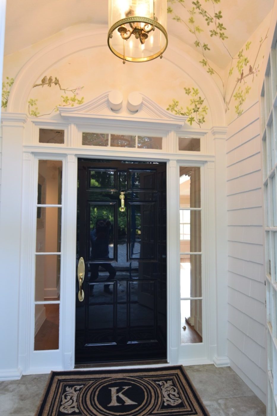 Glossy black front door with sidelights and a transom in a white entry with a branch and bird mural on the arched ceiling