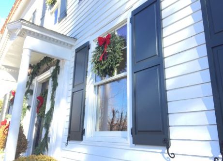 White clapboard house with dark navy shutters, a holiday wreath with a red bow in the window and porch garland