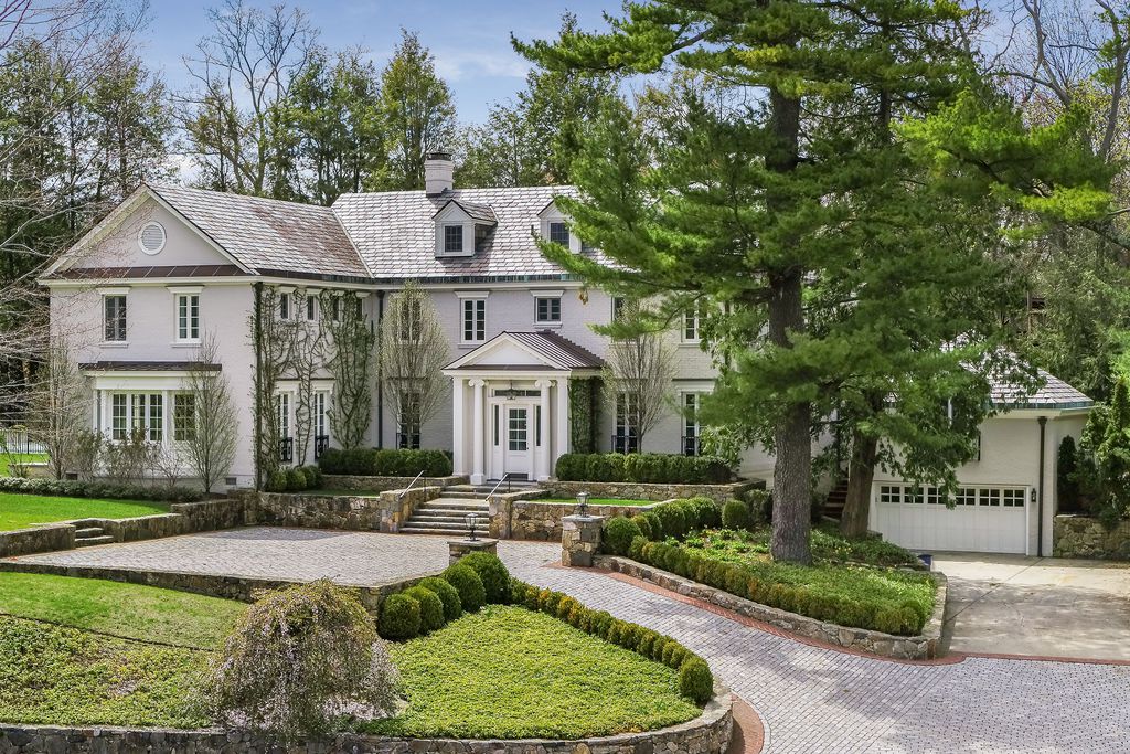 Large white house with a gray slate roof, a circular gravel driveway, boxwood hedges and tall pine trees