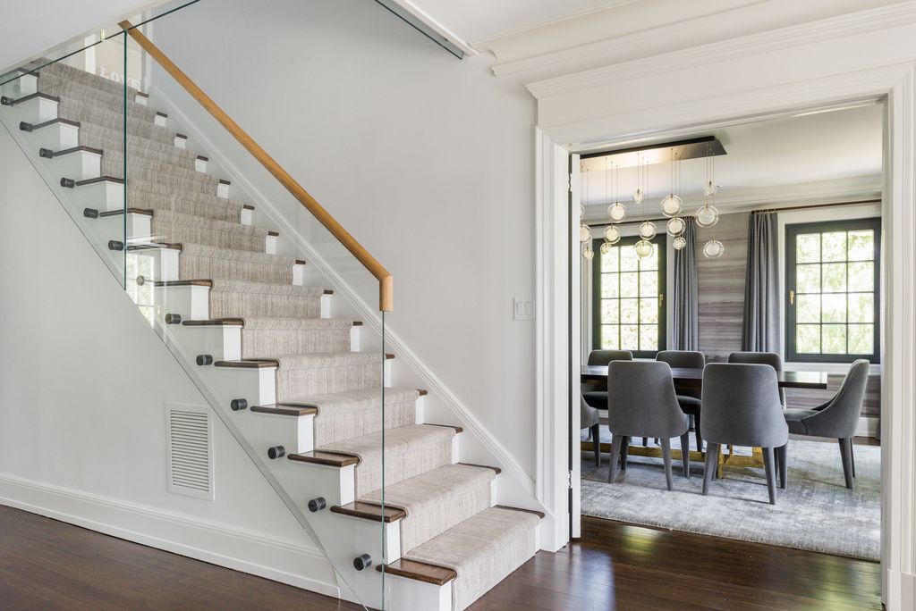 Staircase with a glass railing and carpet runner beside a doorway into a dining room with gray chairs