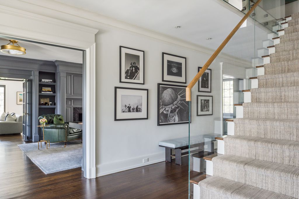 Entry hall with white walls, framed black and white photos, a glass stair railing and a view into a gray paneled den