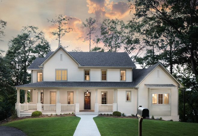 White two-story farmhouse with a dark roof, a wide front porch and lit windows at dusk