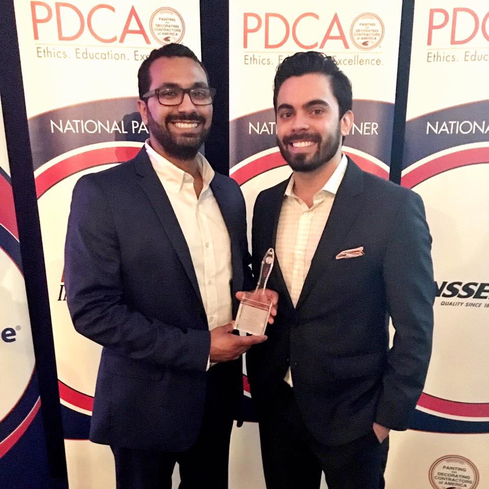 Two team members in suits holding a glass PDCA award trophy in front of a PDCA event backdrop