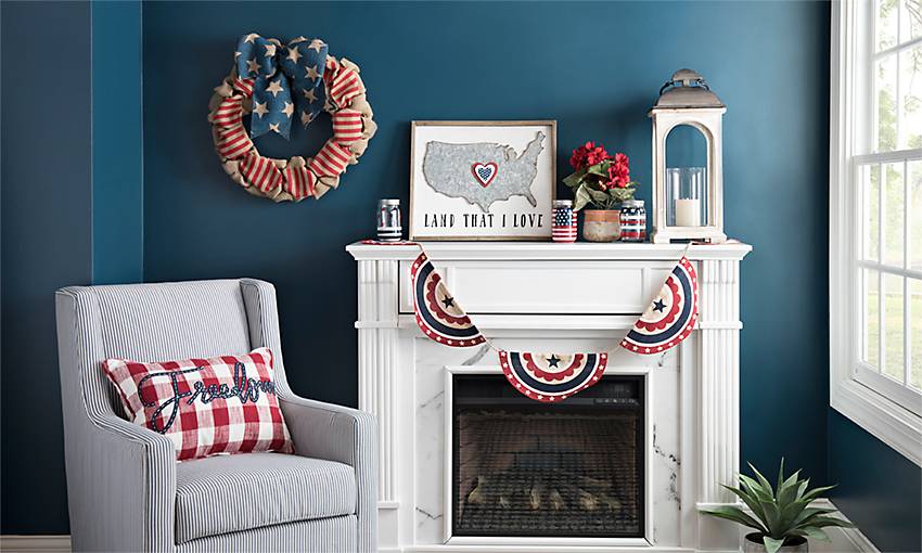 Blue wall with a white fireplace mantel dressed in patriotic bunting, a flag wreath, a lantern and a gray armchair