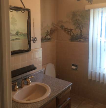 Bathroom with peach wall tile, a painted country scene mural above it, a beige sink and a brass faucet