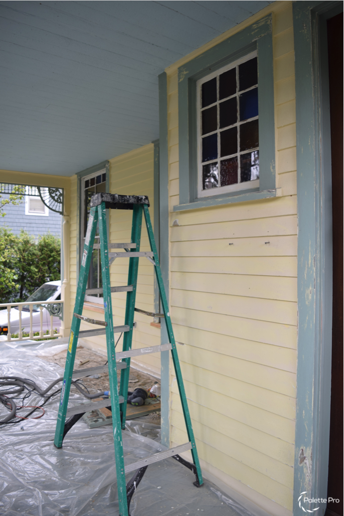 Porch prep with a green stepladder on plastic sheeting beside yellow clapboard and peeling blue-green window trim