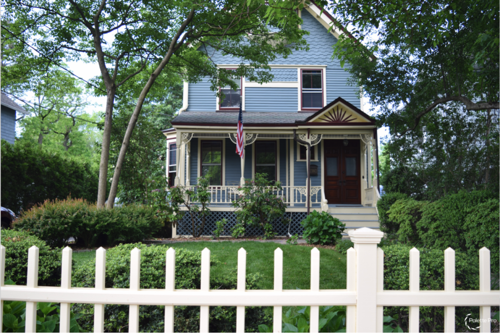 Blue Victorian house with cream trim, a red front door, a decorative porch gable and a white picket fence