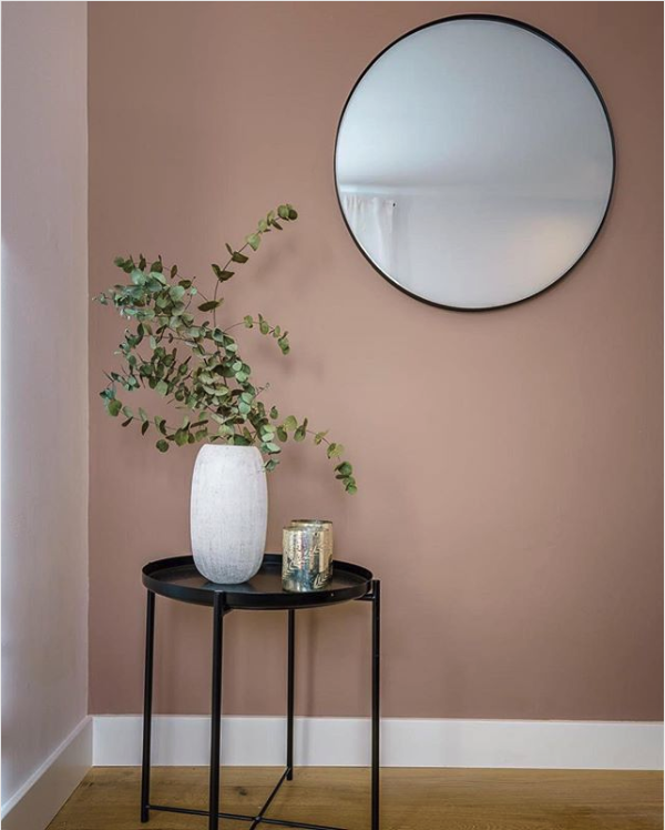 Dusty pink wall with a round black framed mirror above a small black side table with eucalyptus in a white vase