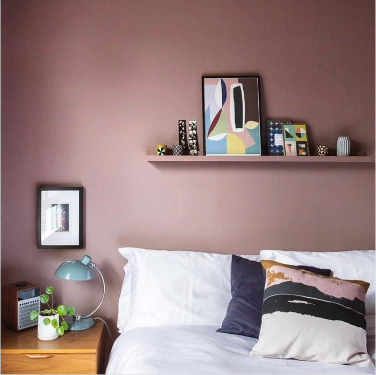 Bedroom with a dusky mauve pink wall, a picture ledge with colorful art, white bedding and a blue desk lamp