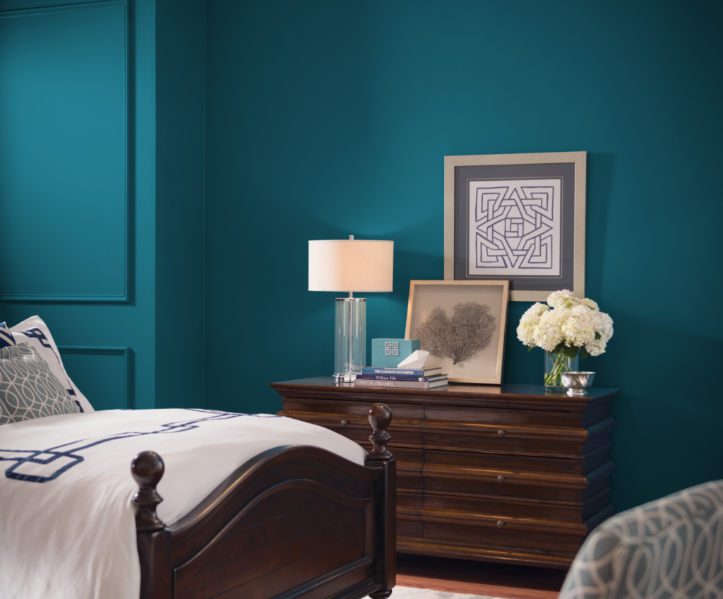Bedroom with deep teal paneled walls, a dark wood bed with white bedding and a dresser with a lamp and flowers