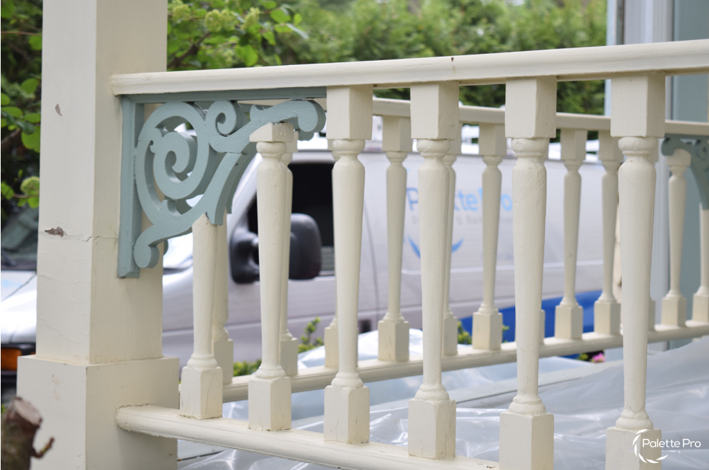 Close-up of cream turned porch balusters and a blue-green scroll bracket with worn paint, plastic sheeting below