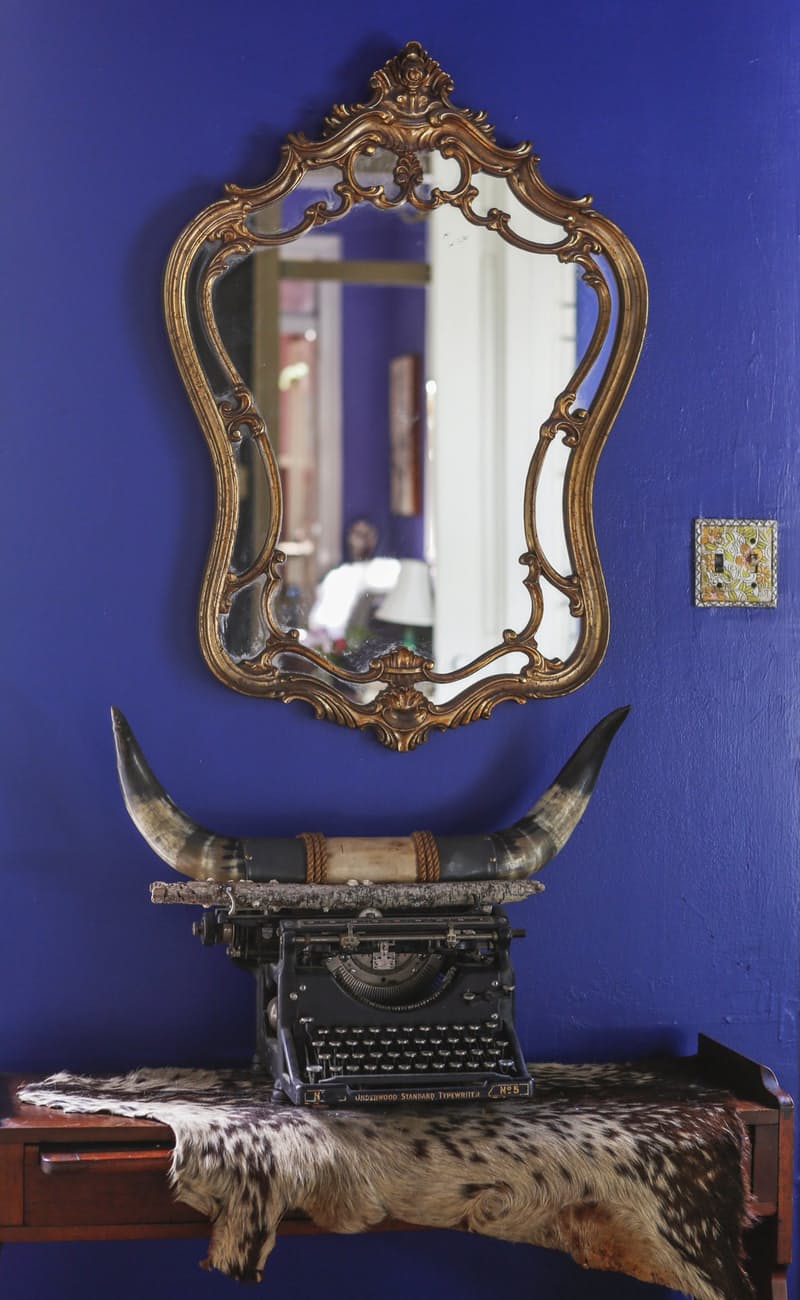 Cobalt blue wall with an ornate gold mirror above a vintage typewriter, mounted horns and a cowhide throw