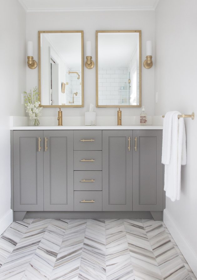 Bathroom with a gray double vanity, brass faucets and pulls, two brass-framed mirrors and a chevron marble floor