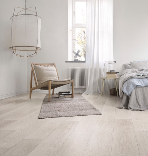 Minimal white bedroom with pale whitewashed wood floors, a wooden lounge chair, a jute rug and gray linen bedding