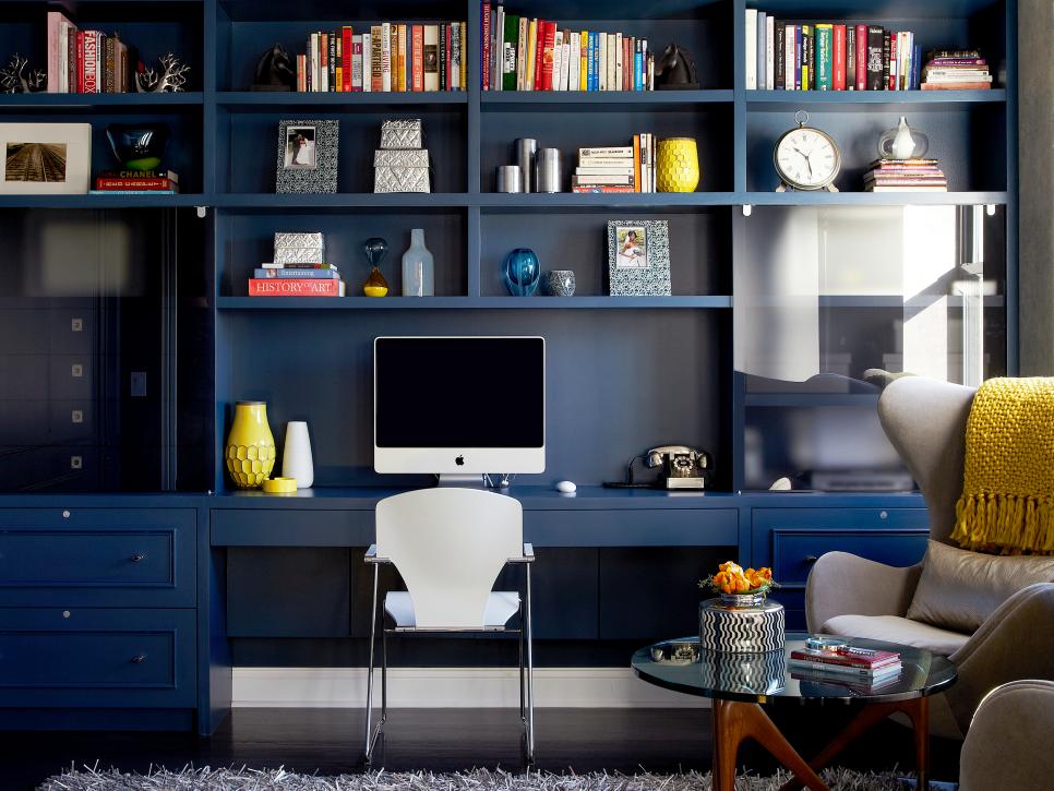 Home office with navy blue built-in bookshelves and desk, a computer, a white chair and yellow accents