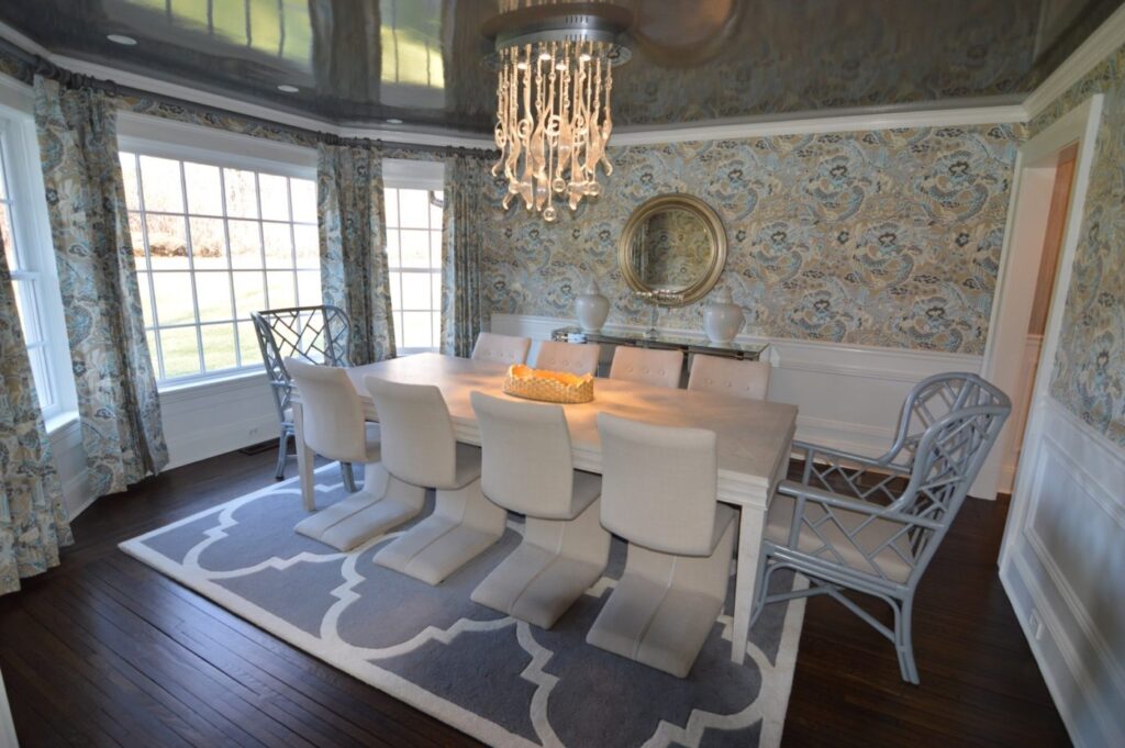 Dining room with blue-gray patterned wallpaper and drapes, a glossy ceiling, a crystal chandelier and white chairs