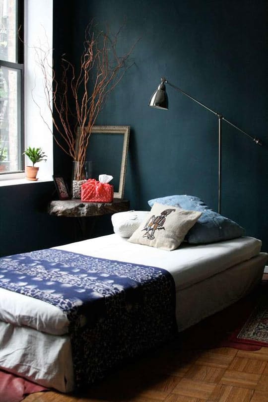 Bedroom with deep teal walls, a low white bed with an indigo throw, a chrome floor lamp and branches in a vase
