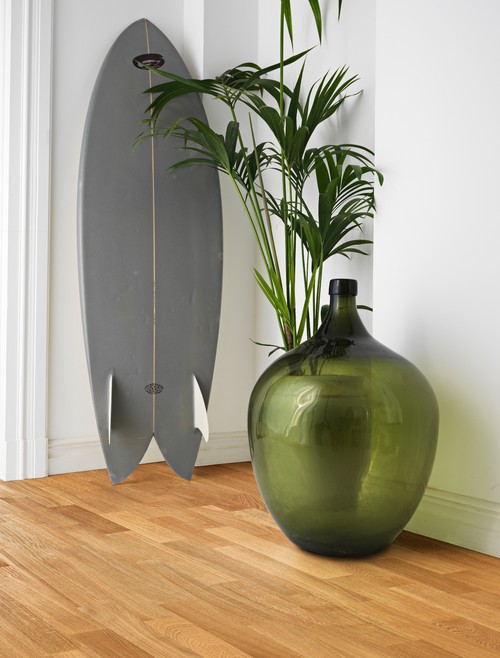 Gray surfboard leaning on a white wall beside a green glass vase with a palm, on a honey oak floor